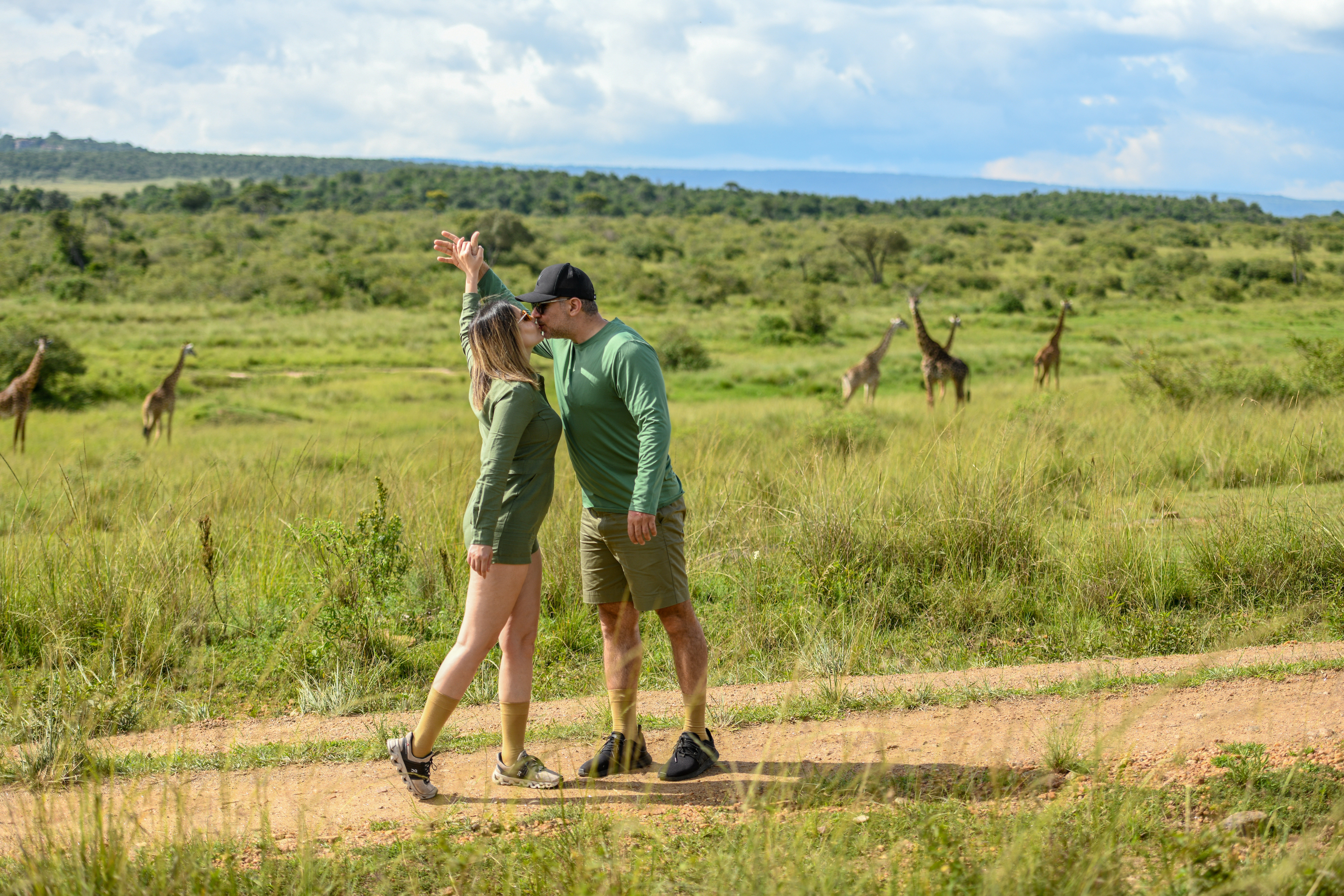 Couple kissing on a bush track with giraffes roaming behind them
